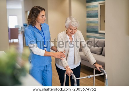 Lovely nurse helping old woman to walk at nursing home with walker. Young nurse helping senior patient using a walking frame to walk in hospital corridor. Caregiver and disabled lady in care facility. Royalty-Free Stock Photo #2003674952