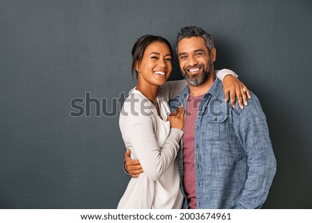 Portrait of happy mid adult couple embracing and looking at camera standing against gray background. Mature indian man in love standing on grey wall while hugging his hispanic woman with copy space. Royalty-Free Stock Photo #2003674961