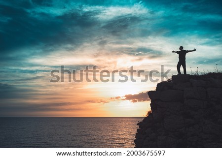 Man silhouette stands on the edge of the abyss and looks the sea with beautiful colorful sky. Royalty-Free Stock Photo #2003675597