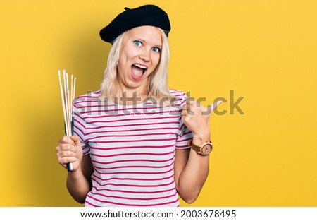 Young caucasian woman holding paintbrushes pointing thumb up to the side smiling happy with open mouth 