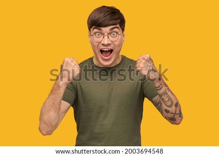 Indoor shot of young male, wears green t-shirt and round stylish spectacles, shouting with widely opened mouth and eyes while watching football game. Isolated over yellow background.