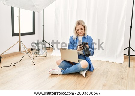 Beautiful blonde woman working as professional photographer at photography studio sitting on the floor checking photos on computer laptop