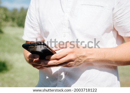 Close-up of male hands browsing the Internet on a mobile phone in nature, outdoors. A man in a white shirt typing on a smartphone in a park on a summer day. Selective focus on hands
