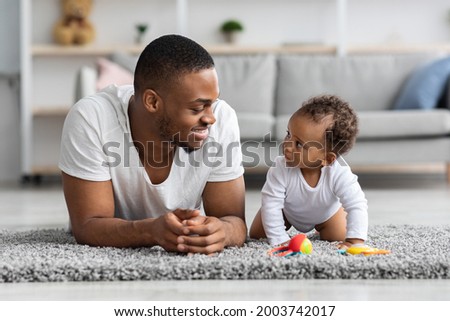 Young Black Dad And His Adorable Infant Child Relaxing Together At Home, African American Father And Cute Newborn Son Lying On Carpet In Living Room, Baby Boy Looking At Daddy With Curiosity Royalty-Free Stock Photo #2003742017