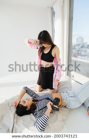 young women taking photo of trendy armenian friend lying on floor on white bedding