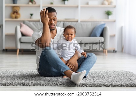 Exhausted Black Young Father Sitting With Crying Newborn Baby On Floor At Home, Desperate African American Dad Can Not Manage Upset Infant Child, Suffering Problems During Paternity Leave Royalty-Free Stock Photo #2003752514