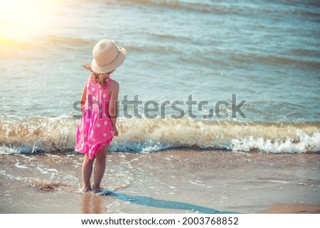 Happy little girl looks at the sea on the beach in summer back to camera. The kid in pink polka-dot dress and straw hat