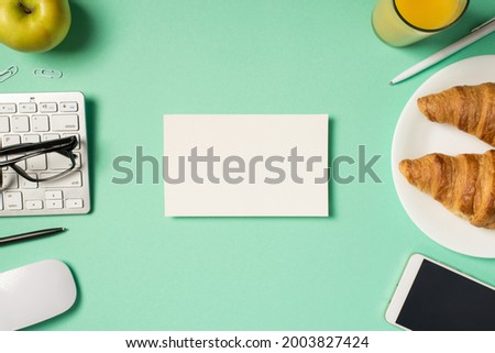 Top view photo of white rectangular card in the middle keyboard glasses mouse pens clips mobile phone apple glass of juice and plate with croissants on isolated turquoise background with blank space