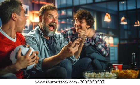 Night at Home: Three Soccer Fans Sitting on a Couch Watch Game on TV, Use Smartphone App to Online Bet, Celebrate Victory when Sports Team Wins. Friends Cheer Eat Snacks, Watch Football Play. Royalty-Free Stock Photo #2003830307
