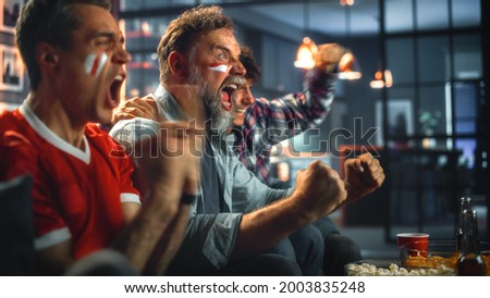 At Home Three Joyfuls Sports Fans with Painted Faces Sitting on a Couch Watch Game on TV, Celebrate Victory when Sports Team Wins Championship. Friends Cheer, Shout. Royalty-Free Stock Photo #2003835248