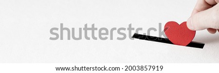 Red heart symbol is put by person's hand into slot of white donation box. Concept of donorship, life saving or charity, sincere devotion to faith. Close-up shot, horizontal banner with copy space Royalty-Free Stock Photo #2003857919