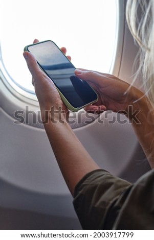 Woman takes pictures on the phone from the airplane window. Hand with a telephone near the porthole. Airplane wing, scenic view. Beautiful cloud, blue sky. Travel by plane, adventure. Air Transport