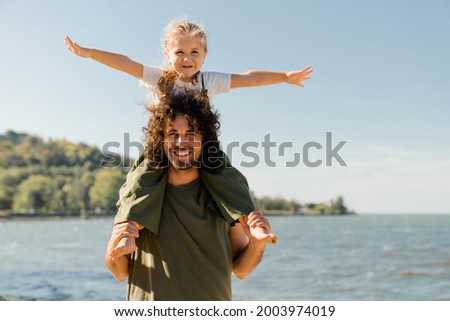 Child daughter sits on her dad shoulders outdoors on a summer beach and looking at camera. Loving father holding little kid child piggyback on weekend walking along lake river