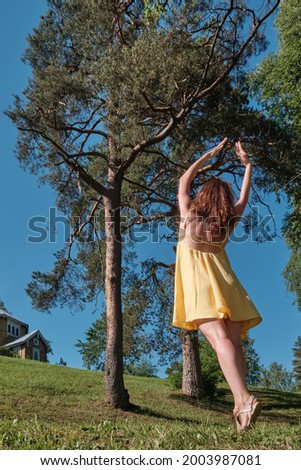 The girl in a yellow dress dances on the lawn near the pines in the sunny weather.
