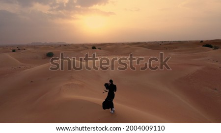 AERIAL. Camera following woman in traditional Emirati dress walking in a desert in strog wind and sunset. Royalty-Free Stock Photo #2004009110