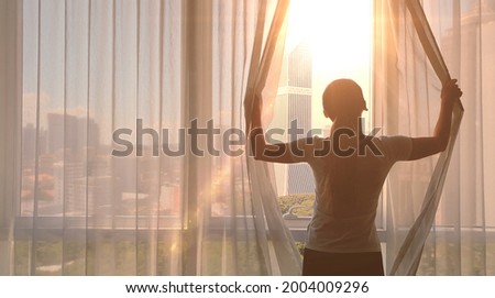 Young woman uncover the big window and looking out her apartment on the city buildings. Sunrise in the city. Royalty-Free Stock Photo #2004009296