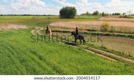 Aerial shot of two horse riders in a beautiful field meadows during the daytime. Horse riders wearing helmet moving across the farm field. Picturesque cloudy sky in the background.