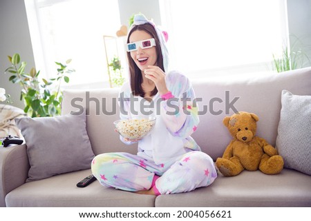 Photo of amazed positive nice funny joyful girl watch 3d film eat pop corn sit sofa wear kigurumi glasses indoors