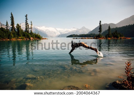 A woman dives into a glacier lake in British Columbia, Canada. Snow capped mountains and trees in the distance. 