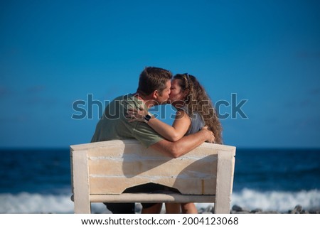 A couple enjoy some time together and a kiss while on vacation. They sit on a cute little bench next to the Caribbean sea