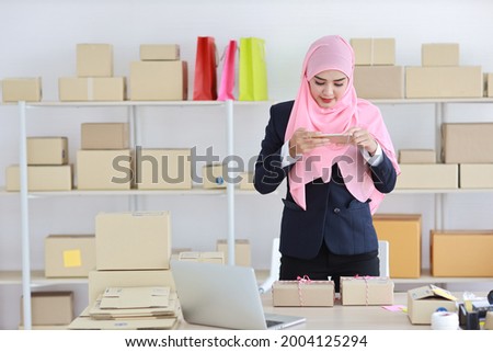 Religious asian muslim woman in blue suit standing and taking picture of package box deliver from mobile phone. Startup small business SME freelance girl work at home with happy smiling face