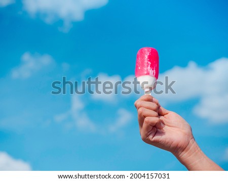 Pink and white popsicle in woman hand. Female hand holding melting pink popsicle on blue sky and cloud background in summer with copy space.