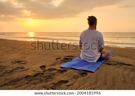 Man practices yoga meditation sitting in padmasana during sunrise. Man sits in lotus position at beach opposite rising sun and its reflection in sea