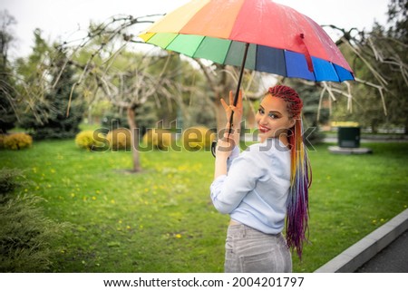 Girl in a bluish shirt with bright makeup and long colored braids. Smiling and holding an umbrella in the colors of the rainbow on the background of a flowered park enjoying the coming spring.