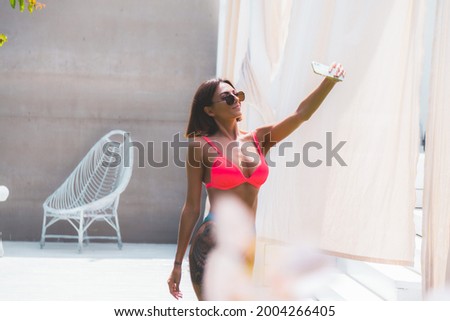 Beautiful tanned fit woman in bikini in backyard with mobile phone take photo selfie, recording video for social media, pool on background