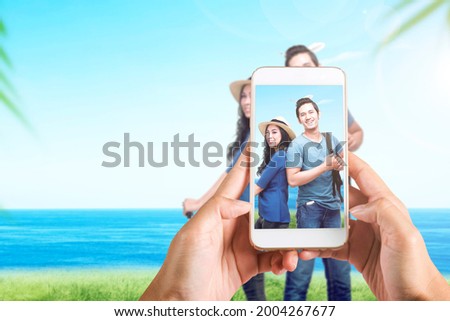 Human hand with the mobile phone taking a picture of Asian couple traveling on the field with seascape background