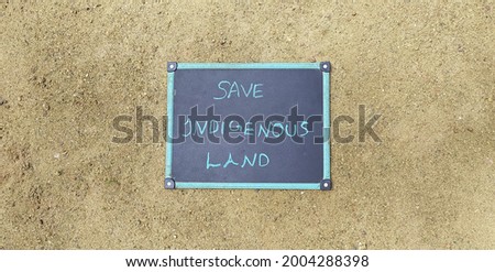 Conceptual protest sign photo of protestors left over isolated black board on dry ground soil of protested area with save indigenous land message written on it. Horizontal closeup flat lay top view. Royalty-Free Stock Photo #2004288398