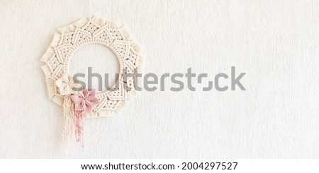 Macrame wreath with cotton flowers on a white decorative plaster wall. Natural cotton thread and rope. Eco decor for home. Creative woman hobby. Copy space. Banner