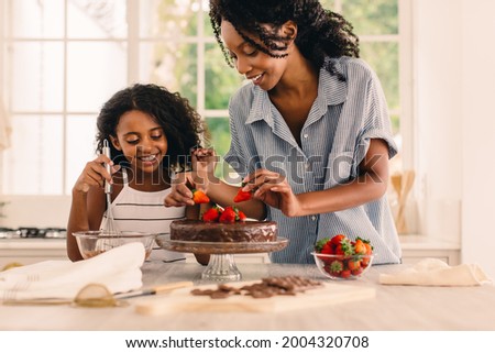 Cute girl standing by her mother in kitchen making cake. Mother and happy daughter garnishing chocolate cake with fresh strawberries in kitchen. Royalty-Free Stock Photo #2004320708