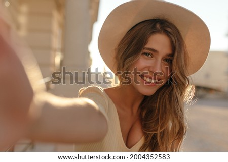 Photo shows close-up of young woman in light hat on blurred background. She has long brown hair and snow-white beautiful smile.