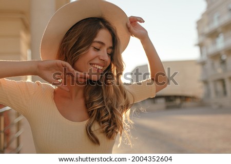 Portrait of cute lady with brown wavy hair in beige hat against background of city. The eyes are closed, and smile is happy and wide. Hands are raised up at level of face.