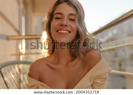 Seductive young woman looking at camera and smiling with teeth while sitting on terrace close-up. The bare and thin curves of collarbones are striking. Royalty-Free Stock Photo #2004352718