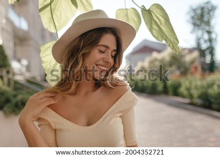 Sunny image of young stylish woman standing on street, in fashionable hat close-up. She has gentle smile and closed eyes. Nice neckline and bare shoulders. Royalty-Free Stock Photo #2004352721