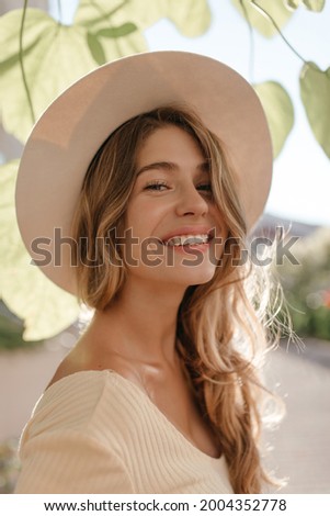 Close-up romantic cute lady smiles broadly and looks into camera. She has wavy blonde hair, beautiful beige hat and blouse.
