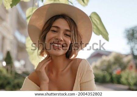 Close-up portrait of lovely white female model with natural make-up expressing good mood. Lovely fair woman in stylish hat holds her chin with her hand. Royalty-Free Stock Photo #2004352802