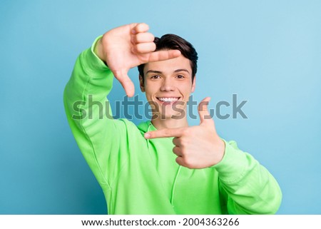 Photo of attractive guy fingers make focus frame make photo wear green hoodie isolated blue color background