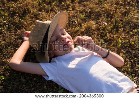 A cute boy with a hat is lying down on the grass and smiling at the sunset.