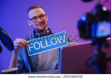 A blogger holds a sign to follow, while recording another video clip. The vlogger wants new viewers to subscribe to it.