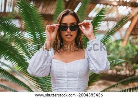 Portrait of a teenager girl touching her sunglasses. She is at the park and there is a palm tree. (Horizontal shot)