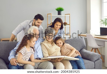 Happy extended family reading together. Young parents, little kids and mature grandparents sitting on sofa at home, enjoying book of good kind educational stories or looking through family photo album Royalty-Free Stock Photo #2004385925