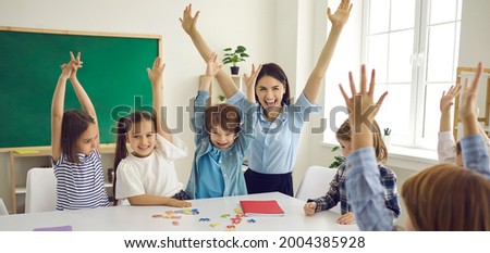 Happy children and educator raising hands up sitting at table in modern classroom. Website header with group portrait of cheerful school teacher and first grade students having fun activities in class Royalty-Free Stock Photo #2004385928