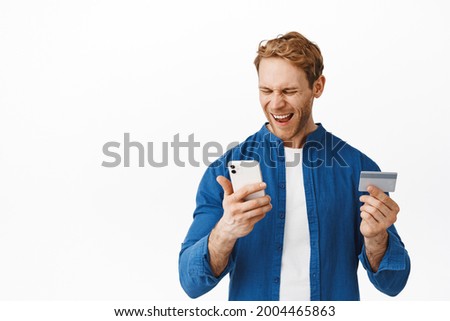 Satisfied and happy redhead guy looking at his mobile phone and shouting yes, holding credit card, winning money, receive cashback, standing over white background