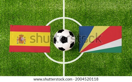 Top view soccer ball with Spain vs. Seychelles flags match on green football field.