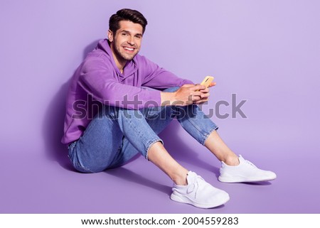 Full length body size photo man smiling typing message on smartphone sitting isolated pastel violet color background