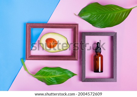 Glass dropper bottle and avocado inside picture frames against the pink and blue geometric background. Ingredient of the beauty product. Flat lay