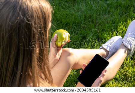 Young woman eats green apple and uses her smartphone, woman sits on grass on a sunny day and uses the internet. Woman eats fruit and check email, messages.